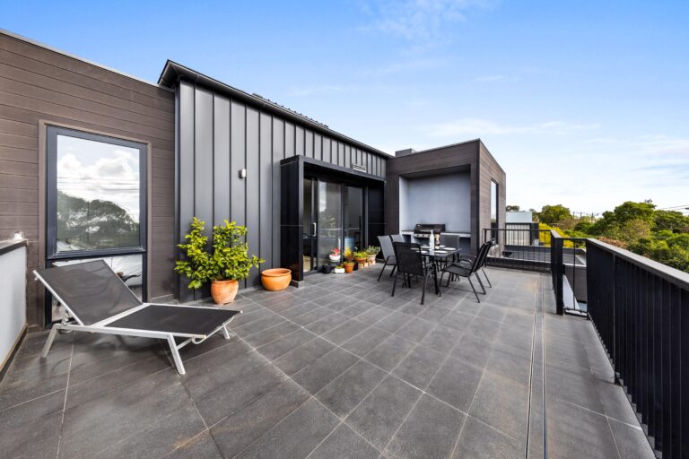 Spacious modern rooftop terrace with dark tiled flooring, outdoor dining set, potted plants, and a sun lounger, featuring sleek black and grey exterior walls and a view of surrounding greenery under a clear blue sky.